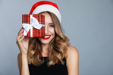 Happy young woman wearing Santa hat with Christmas gift on grey background