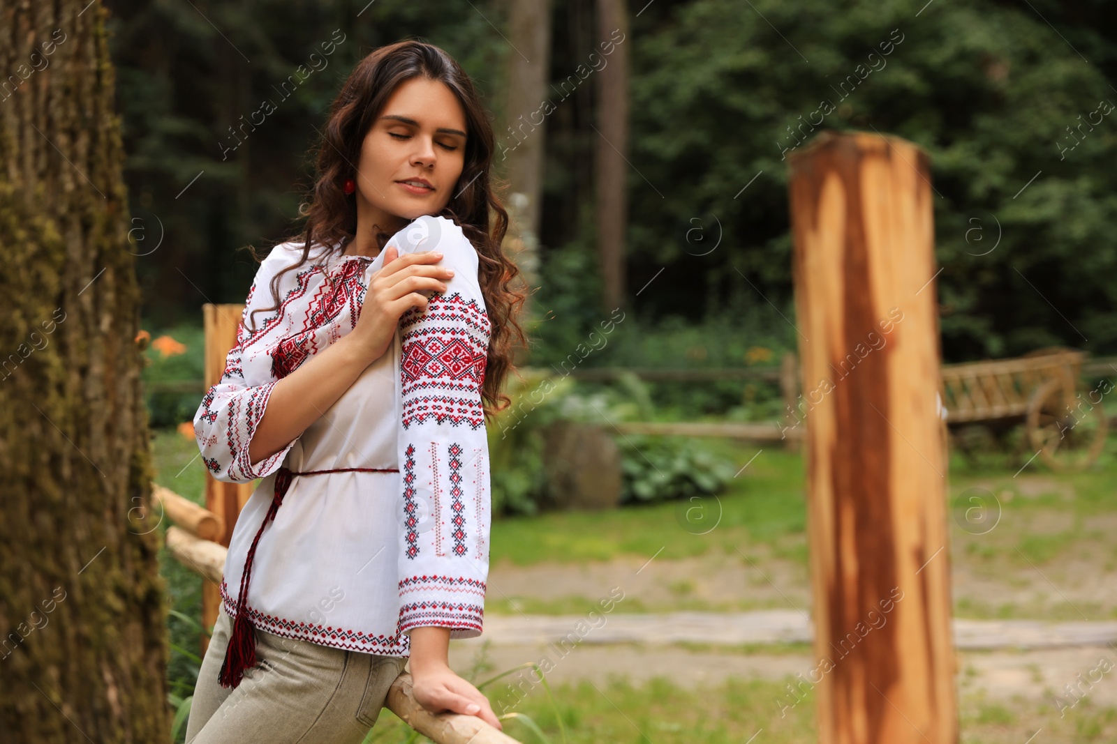 Photo of Beautiful woman wearing embroidered shirt near wooden railing in countryside, space for text. Ukrainian national clothes
