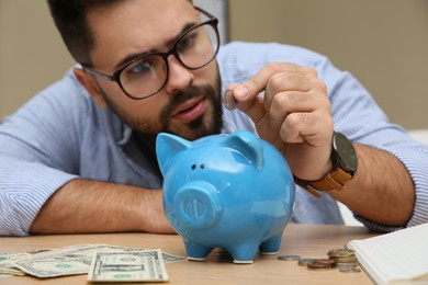 Photo of Young man putting coin into piggy bank at table indoors, focus on hand. Money savings