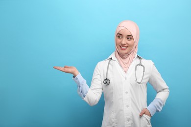 Muslim woman in hijab and medical uniform pointing at something on light blue background