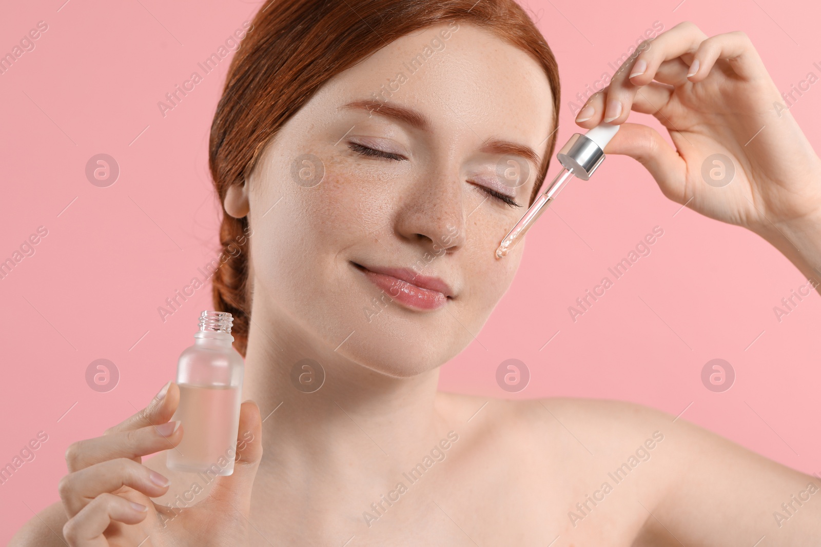 Photo of Beautiful woman with freckles applying cosmetic serum onto her face on pink background, closeup