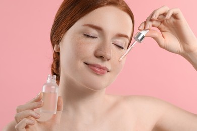 Beautiful woman with freckles applying cosmetic serum onto her face on pink background, closeup