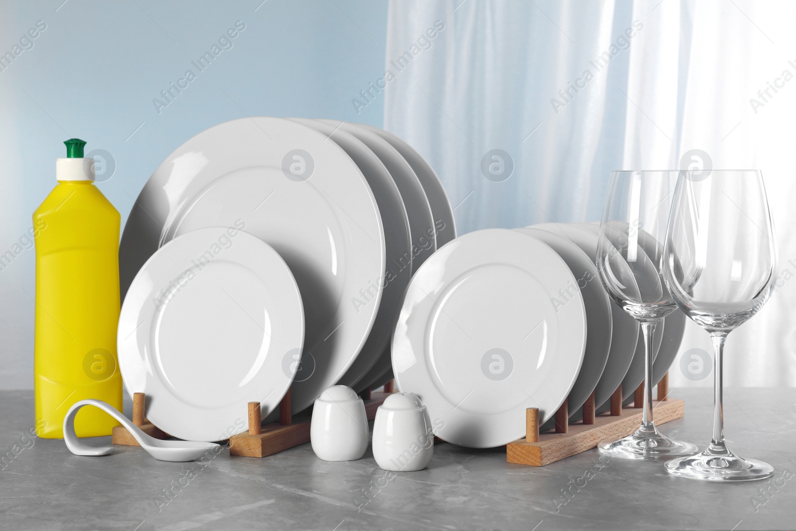 Photo of Glasses, clean dishware and bottle of detergent on grey marble table