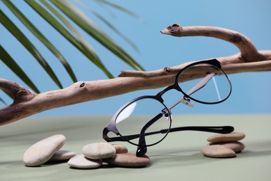 Photo of Stylish pair of glasses with black frame, stones and wooden snag on olive table against blurred background