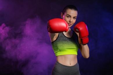 Portrait of beautiful woman wearing boxing gloves training in color lights and smoke on black background. Space for text