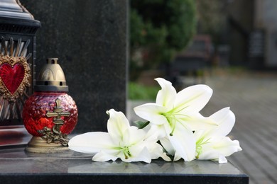 Photo of White lilies and grave light on grey granite tombstone outdoors. Funeral ceremony