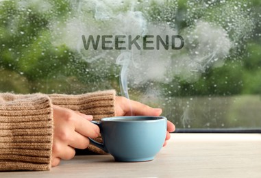 Image of Happy Weekend. Woman with cup of hot drink at wooden table near window on rainy day, closeup