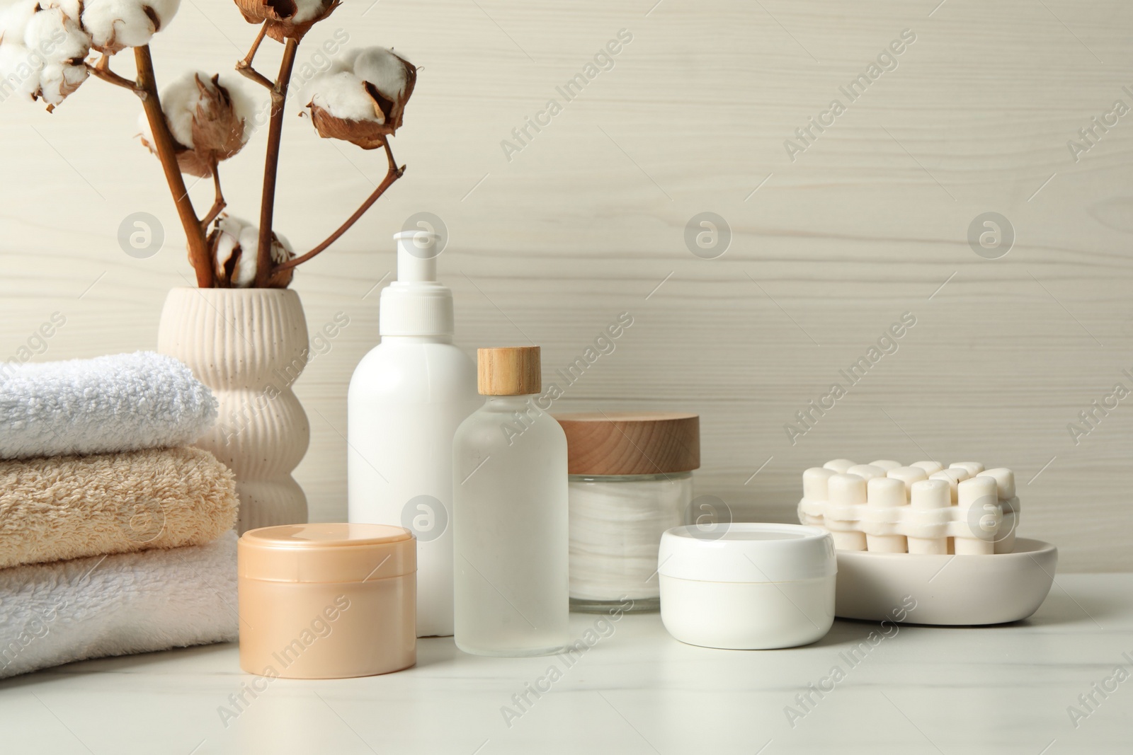 Photo of Bath accessories. Personal care products and cotton flowers in vase on white marble table near light wooden wall