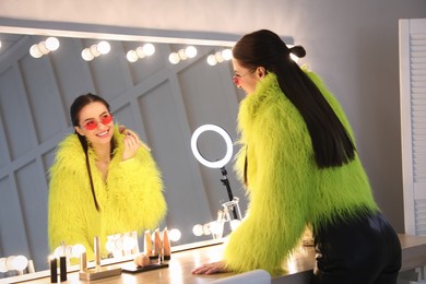 Photo of Young woman applying make up near illuminated mirror indoors