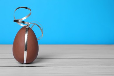 Photo of Delicious chocolate egg with silver ribbon on white wooden table against light blue background, closeup. Space for text