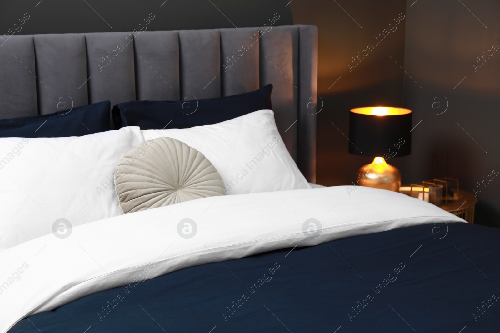 Photo of Comfortable bed with cushions, lamp and different decor on bedside table in room. Stylish interior