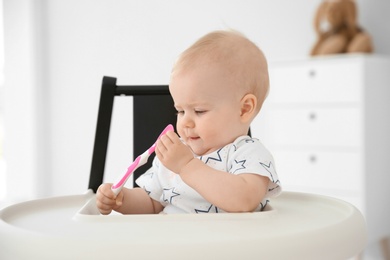 Photo of Cute little baby with spoon in high chair at home