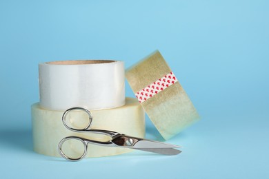 Photo of Rolls of adhesive tape and scissors on light blue background. Space for text