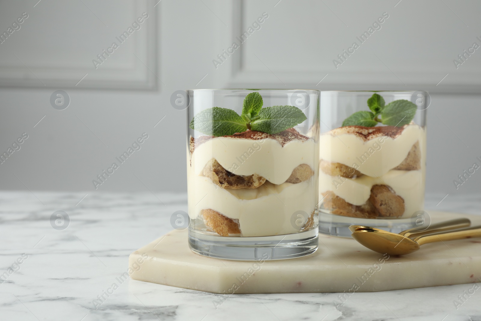 Photo of Delicious tiramisu in glasses, mint leaves and spoons on white marble table, space for text