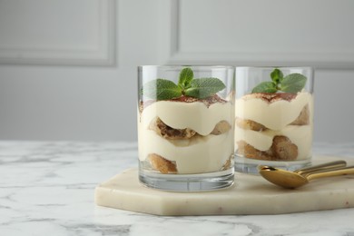 Delicious tiramisu in glasses, mint leaves and spoons on white marble table, space for text