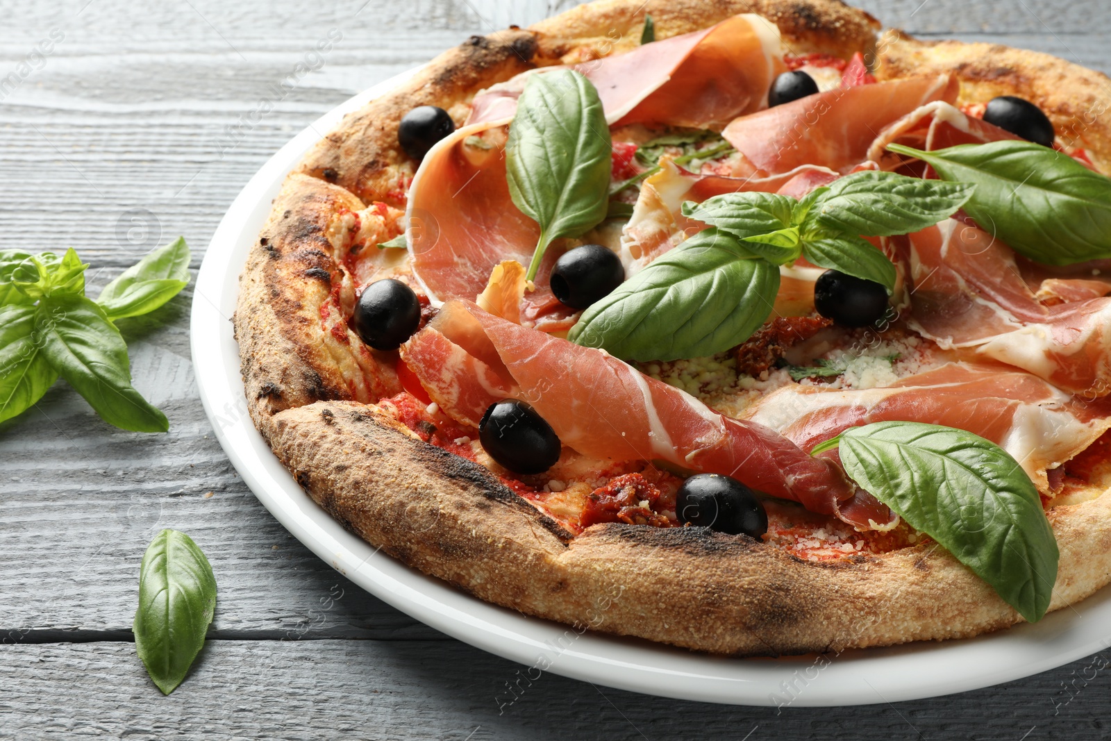 Photo of Tasty pizza with cured ham, olives, sun-dried tomato and basil on gray wooden table, closeup