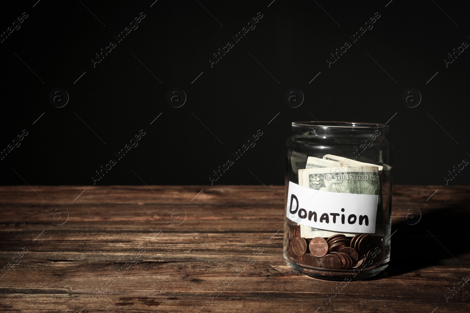 Photo of Glass jar with money and label DONATION on table against black background. Space for text