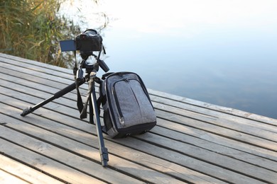 Photo of Tripod with modern camera and backpack on wooden pier near water. Professional photography