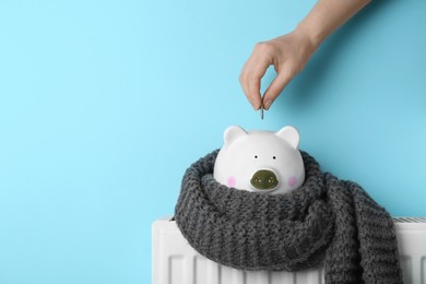 Photo of Woman putting coin into piggy bank on heating radiator against light blue background, closeup. Space for text