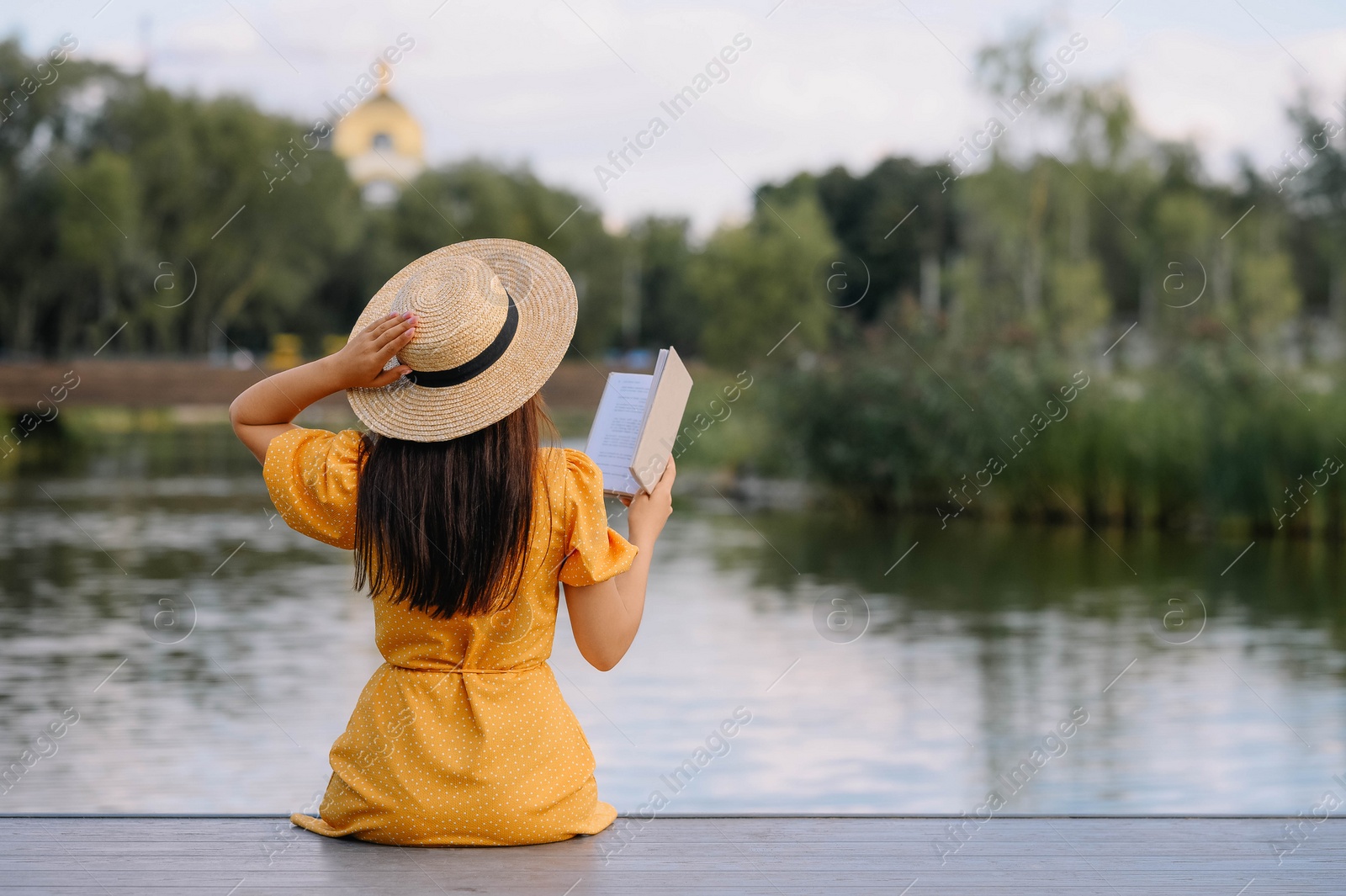 Photo of Woman reading book on pier near lake, back view