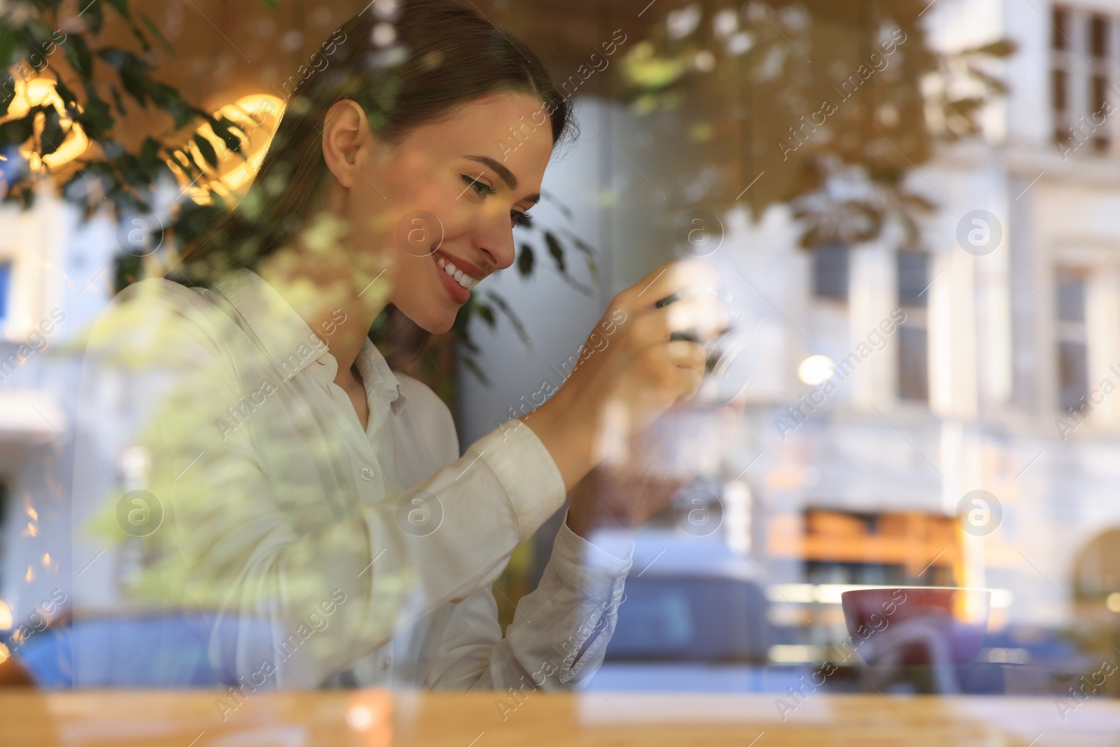 Photo of Young woman taking photo of cup with coffee at cafe, view through window. Creative hobby