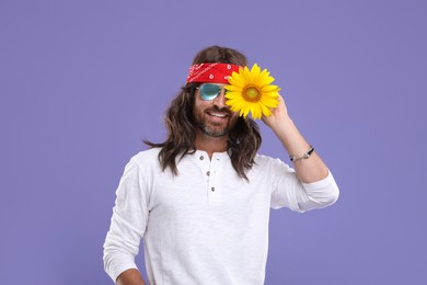 Photo of Stylish hippie man covering eye with sunflower on violet background