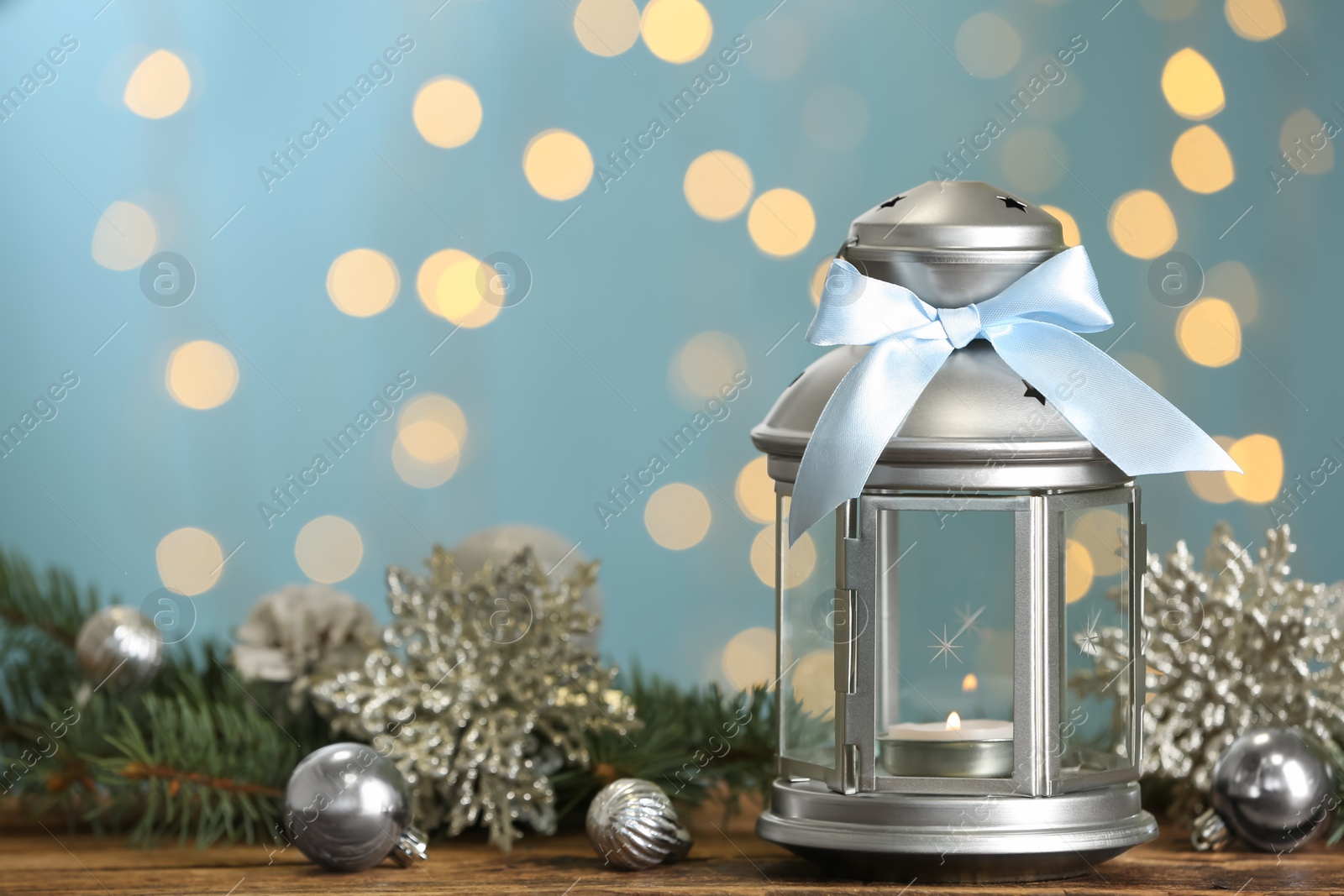 Photo of Christmas lantern with burning candle and festive ornaments on wooden table against blurred lights. Space for text