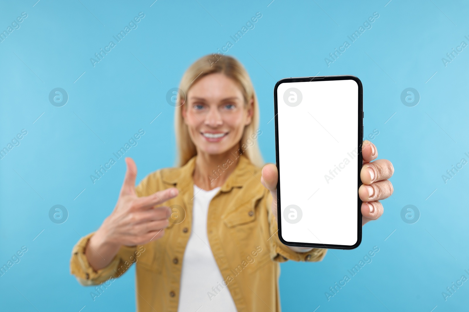 Photo of Happy woman holding smartphone and pointing at blank screen on light blue background, selective focus