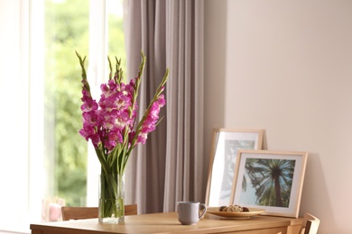 Vase with beautiful pink gladiolus flowers, pictures and cup on wooden table in room, space for text