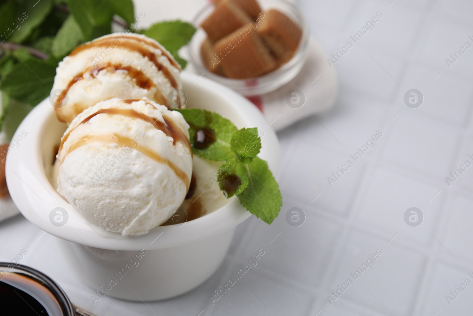 Photo of Scoops of tasty ice cream with mint leaves and caramel sauce on white tiled table, closeup. Space for text
