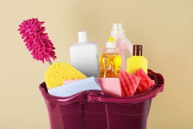 Photo of Bucket with different cleaning supplies against beige background, closeup