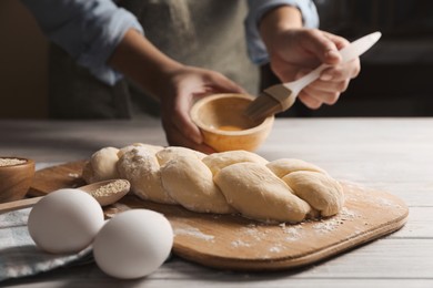 Photo of Woman spreading egg yolk onto raw braided bread at white wooden table, closeup. Traditional Shabbat challah
