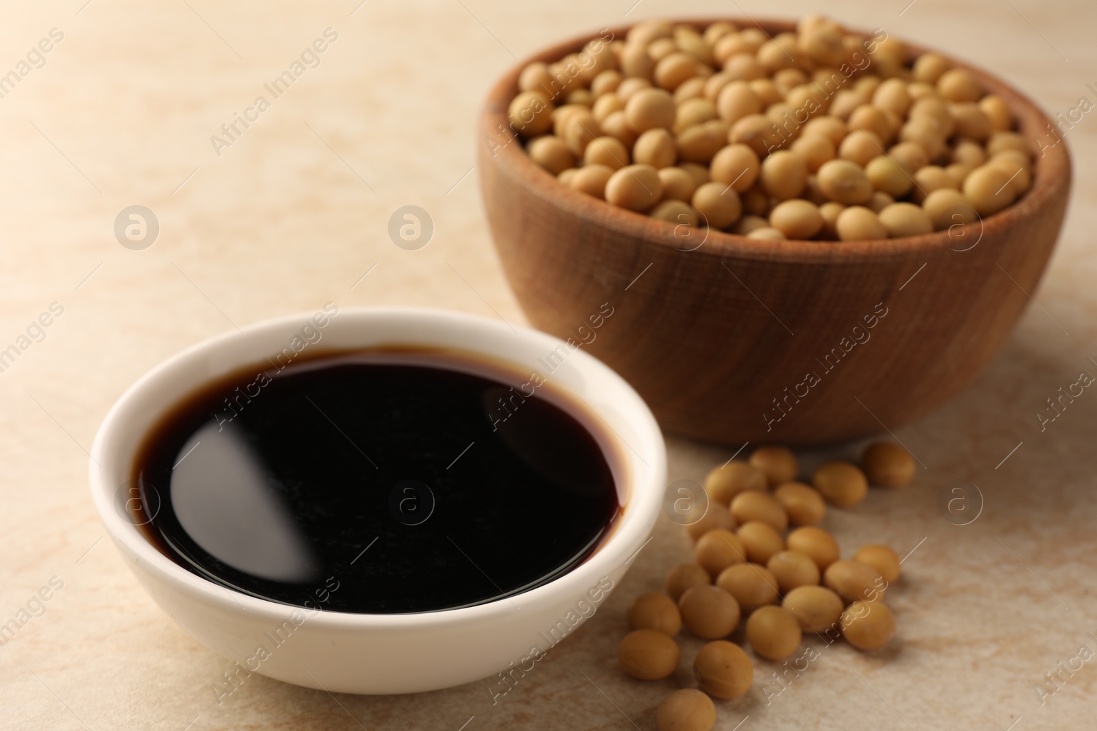 Photo of Soy sauce in bowl and soybeans on beige table, closeup