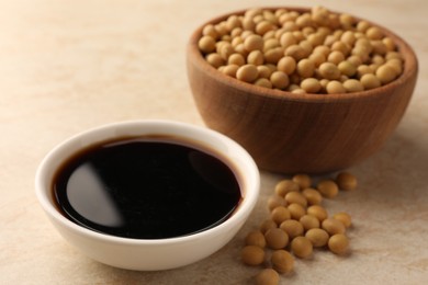 Soy sauce in bowl and soybeans on beige table, closeup