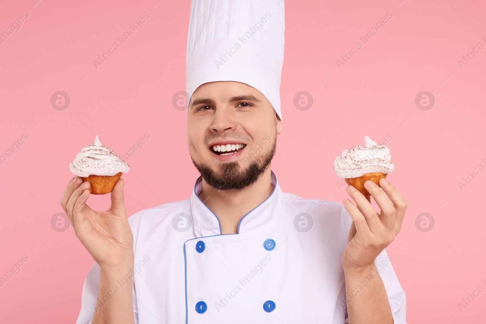 Photo of Happy professional confectioner in uniform holding delicious cupcakes on pink background