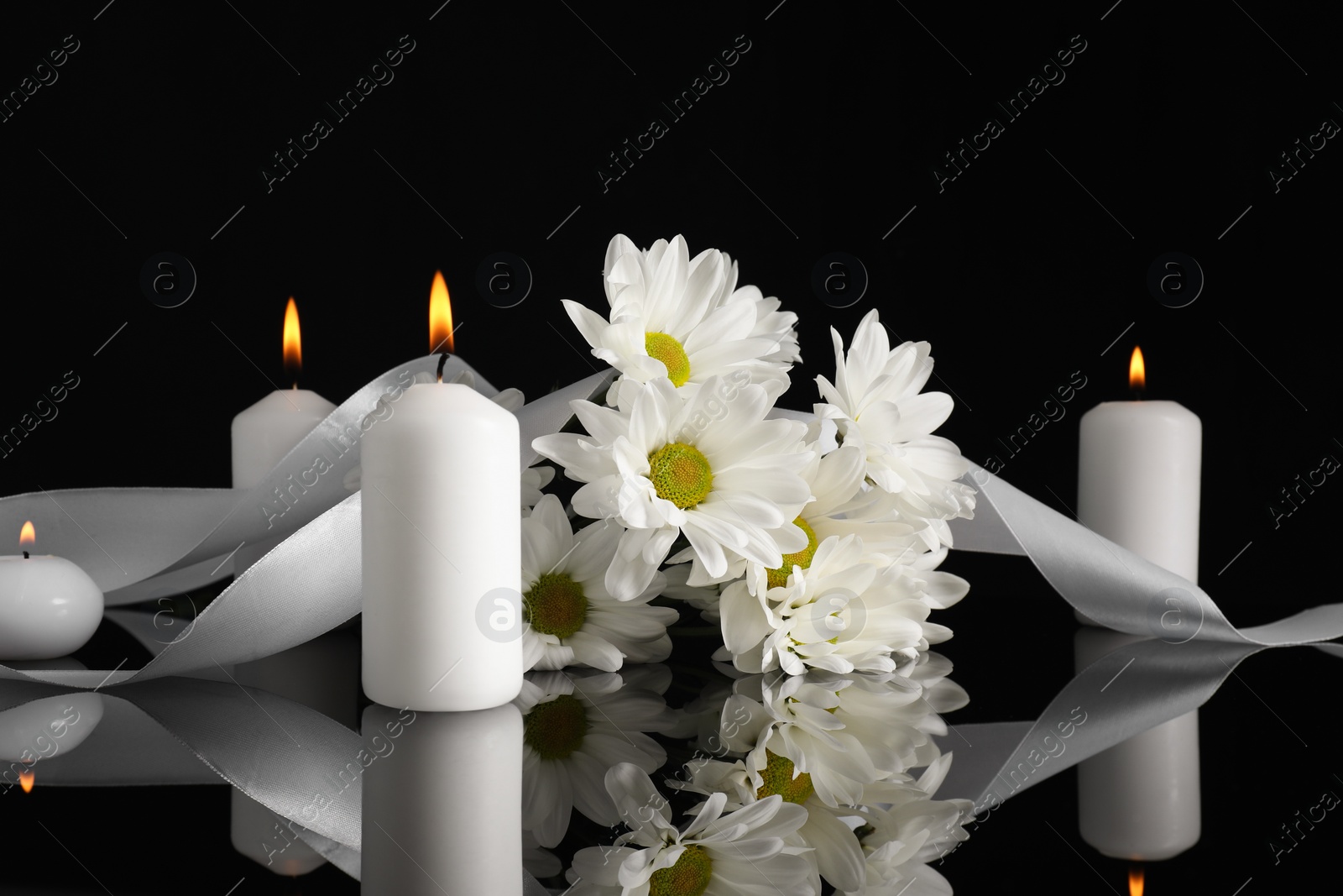 Photo of White chrysanthemum flowers and burning candles on black mirror surface in darkness. Funeral symbols