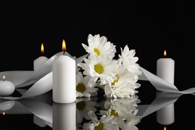 Photo of White chrysanthemum flowers and burning candles on black mirror surface in darkness. Funeral symbols