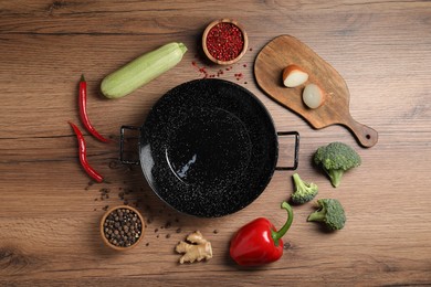 Empty iron wok surrounded by raw ingredients on wooden table, flat lay