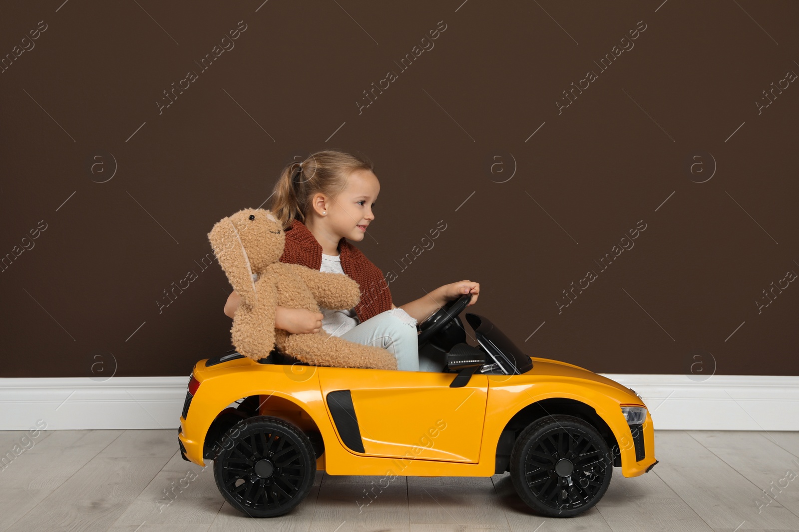 Photo of Cute little girl with toy bunny driving children's car near brown wall indoors