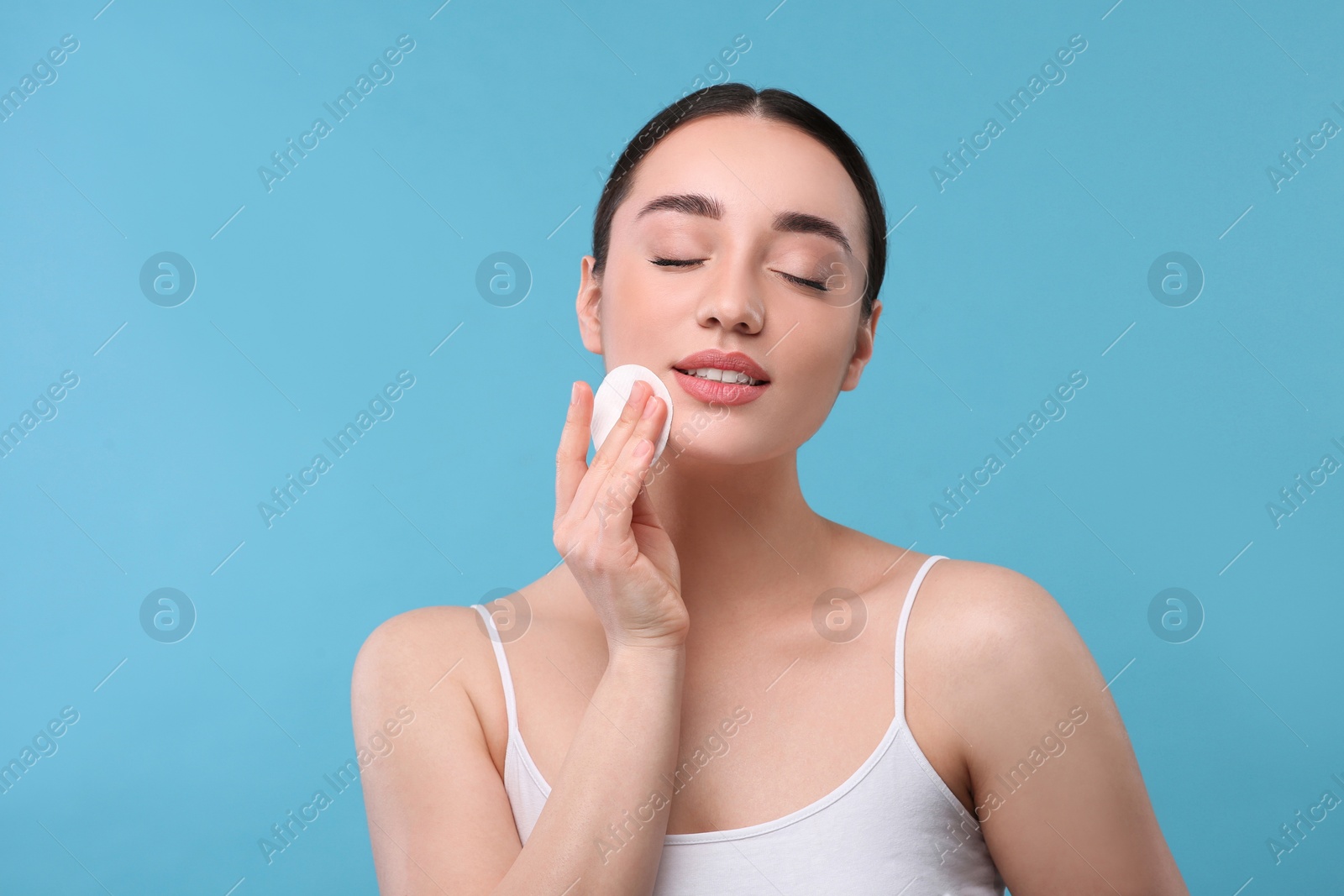 Photo of Beautiful woman removing makeup with cotton pad on light blue background