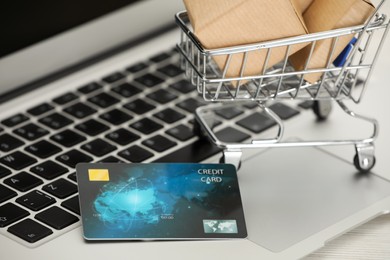 Photo of Online payment concept. Small shopping cart with bank card, boxes on laptop, closeup