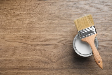 Open can with white paint and brush on wooden background, top view. Space for text