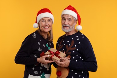 Senior couple in Christmas sweaters and Santa hats holding gift on orange background