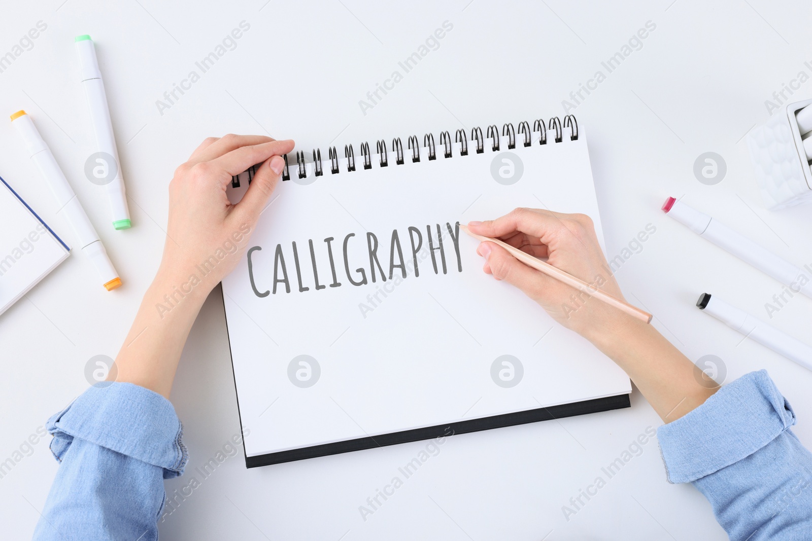 Image of Woman writing word Calligraphy in notebook at white table, top view