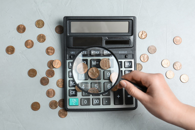 Woman looking through magnifying glass at calculator with coins on light grey table, flat lay. Search concept