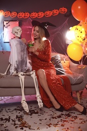 Photo of Woman in witch hat sitting near skeleton on sofa indoors. Fun Halloween party