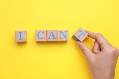 Photo of Motivation concept. Woman changing phrase from I Can't into I Can by removing wooden cube with letter T on yellow background, top view