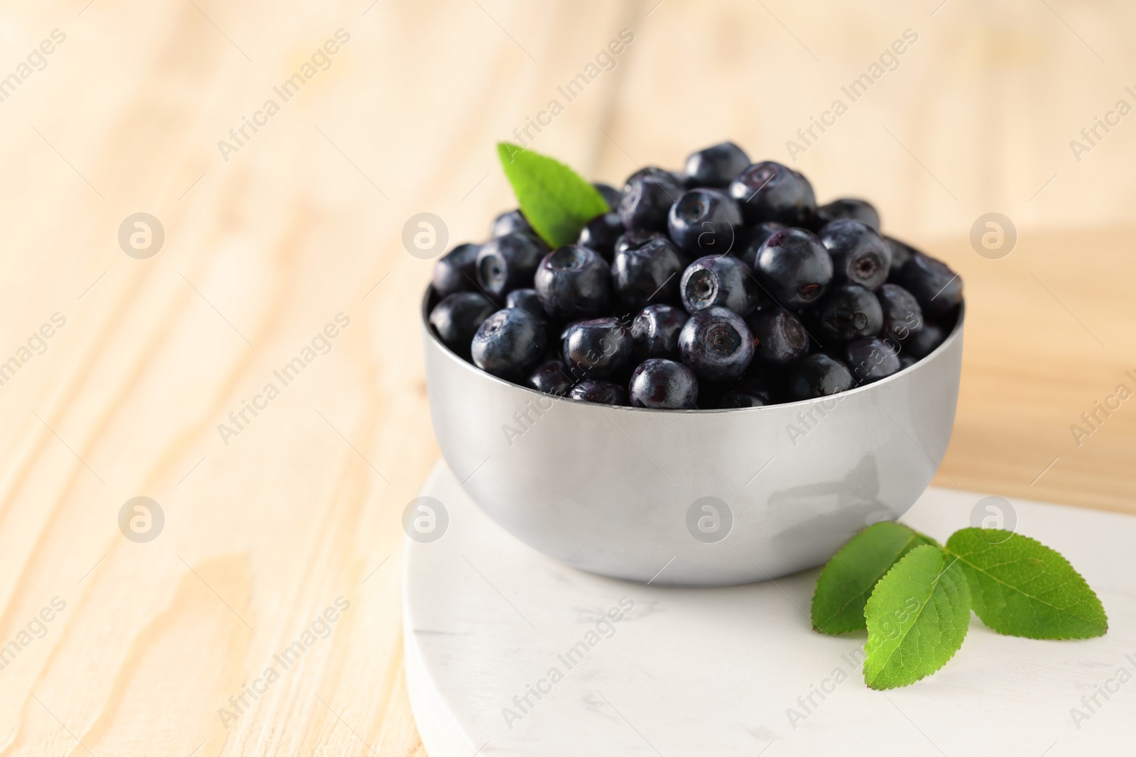 Photo of Bowl of tasty fresh bilberries and green leaves on light table, closeup. Space for text