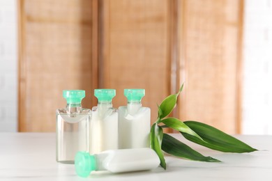 Photo of Mini bottles of cosmetic products and green branch on white table against blurred background