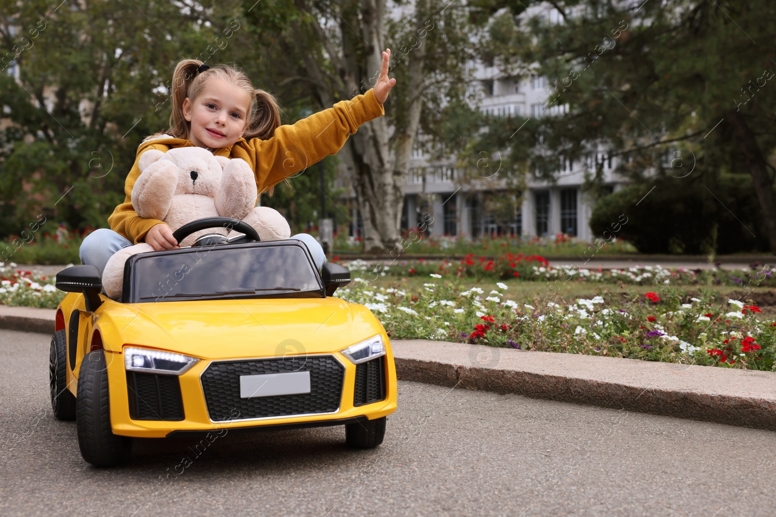 Photo of Cute little girl with toy bear driving children's car in park. Space for text